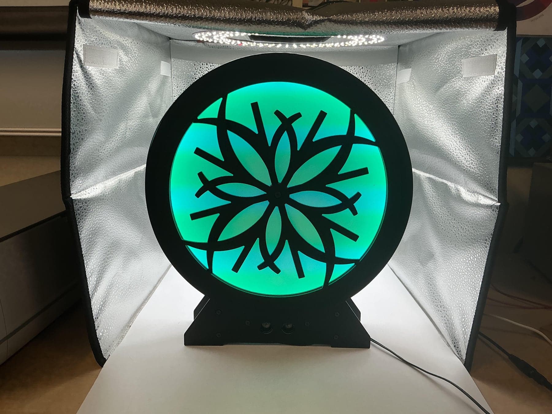The finished kinetic sculpture lit in a photo booth: a black moiré disk in front of a glowing teal illumination plane.