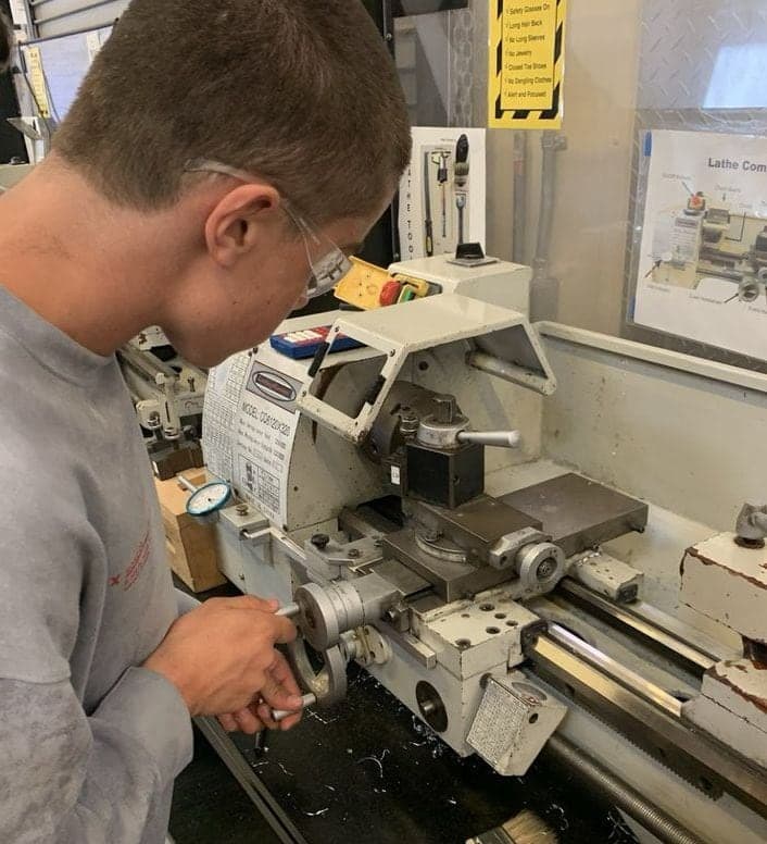 Student in safety glasses turning the cross-slide handwheel of a benchtop metal lathe, with the tool post and chuck guard in front of him.