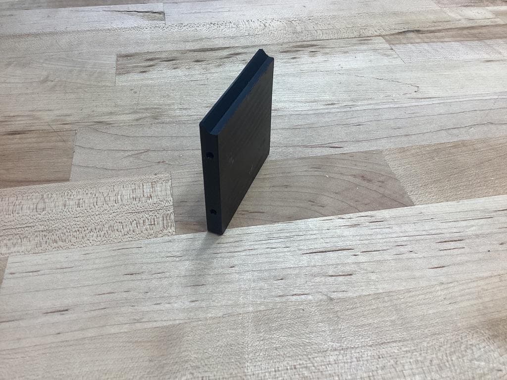 A machined black base plate standing on edge on a maple bench.