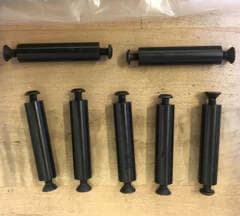 Seven machined black axles laid out on a wooden surface, two horizontal and five vertical, each with a small mushroom-shaped head at one end and a flared countersunk head at the other.