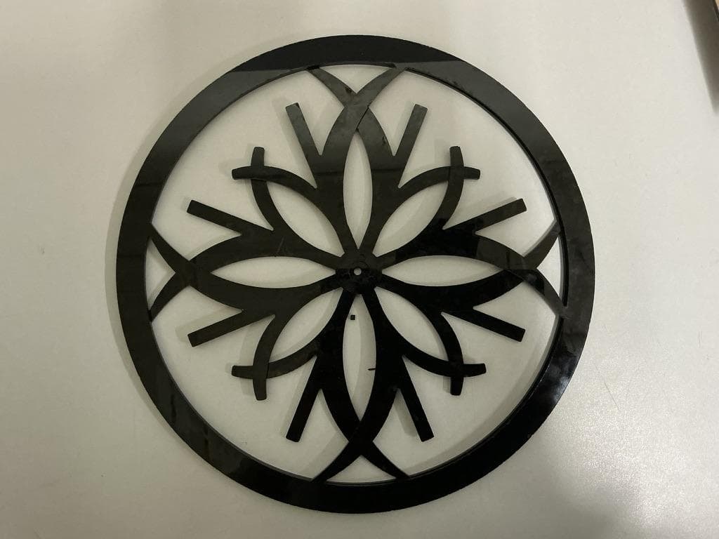 Laser-cut black moiré disk photographed flat on a pale surface: a solid outer ring around interlocking curved petal shapes radiating from a small central hole.