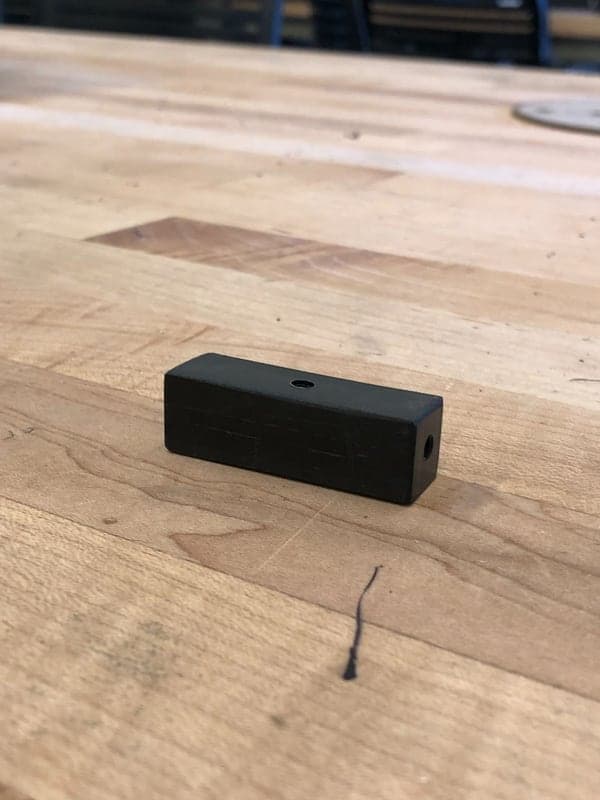 Rectangular black plastic block lying on a wooden workbench, with a small hole in the middle of its top face and another in its end.