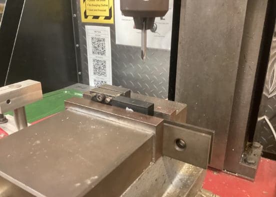 Milling machine spindle holding a pointed drill above a vise that clamps a small black rectangular block against a stop.