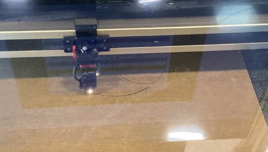Laser cutter head over a brown sheet, seen through a reflective lid, with thin dark curved cut lines forming part of a disk pattern.