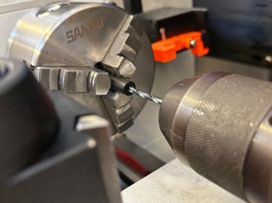 Close-up of a lathe: a twist drill in a tailstock chuck entering the end of a short black rod gripped in a three-jaw chuck.
