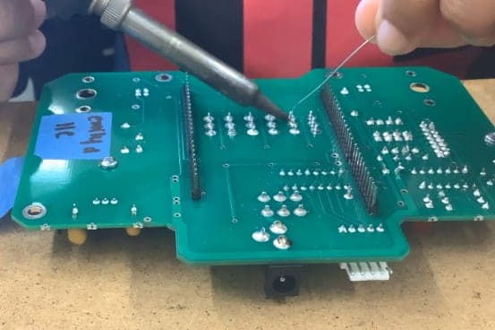 Hands holding a soldering iron and solder wire over a green circuit board with rows of silver solder joints and two long pin headers.