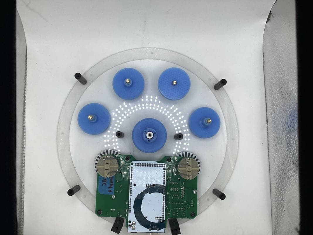 Top-down view of a clear circular plate with five blue pulleys, black standoffs, and a green circuit board holding an Arduino and two motors.