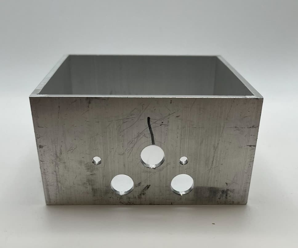Raw machined aluminum base box, open at the top, with holes drilled in its front face.