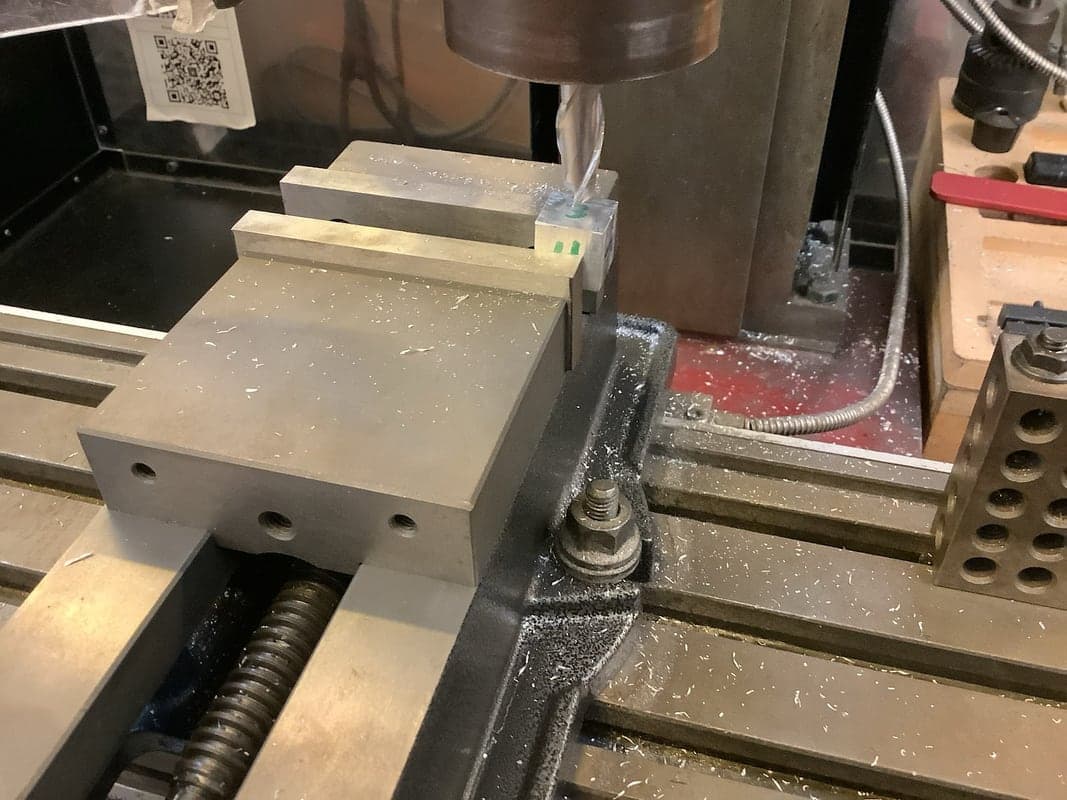 End mill descending onto a small aluminum block held in a machine vise on a mill table, with chips scattered and a 1-2-3 block nearby.