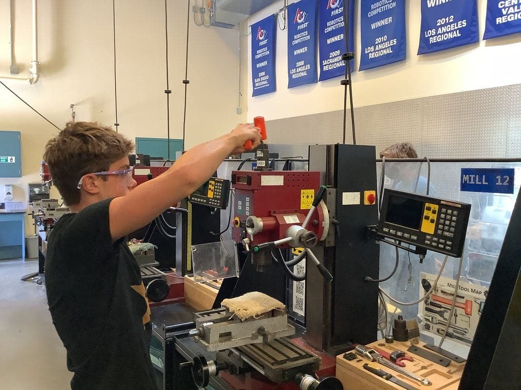 Student in safety glasses raising an orange dead-blow hammer over the drawbar of a red benchtop mill with a digital readout.