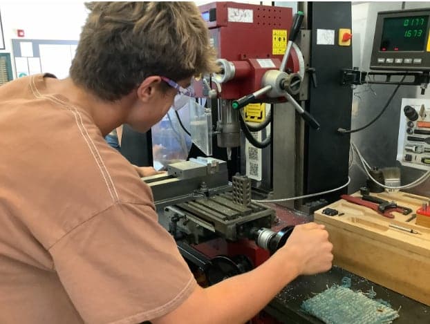 Student in safety glasses cranking the table handwheel of a red benchtop mill, with a vise on the table and a digital readout above.