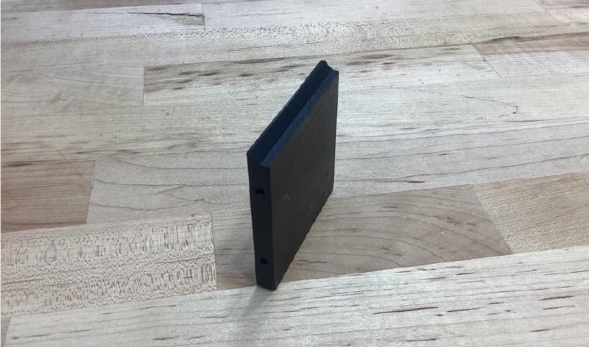 A black machined plate standing on its edge on a maple workbench, showing a stepped ledge along its top edge and two drilled holes in its side.