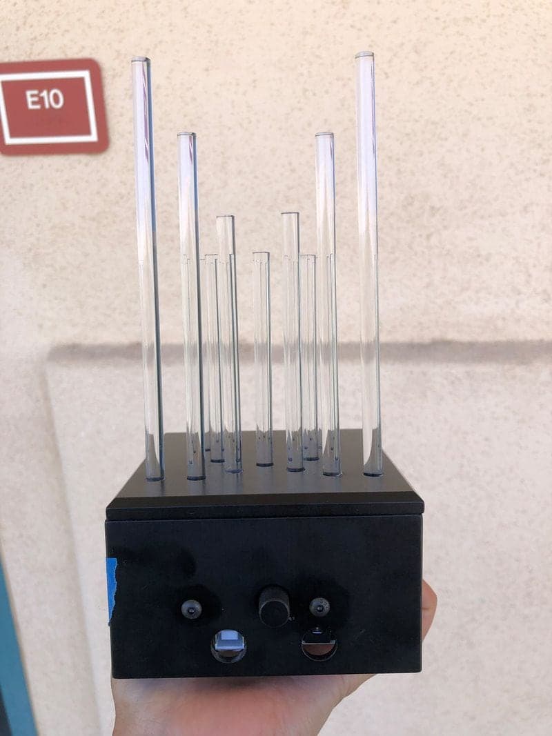 Hand holding a black box with clear acrylic rods of varying heights rising from its top and a black knob and two port openings on its front face.