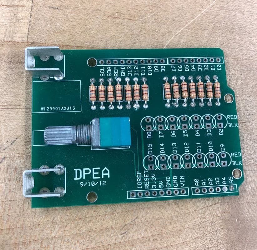 Green DPEA circuit board on a wooden bench with two rows of tan resistors, a teal potentiometer with a metal shaft, and two metal brackets.
