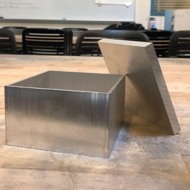 Open square aluminum tube section and a flat aluminum plate leaning beside it on a wooden workbench in a classroom.