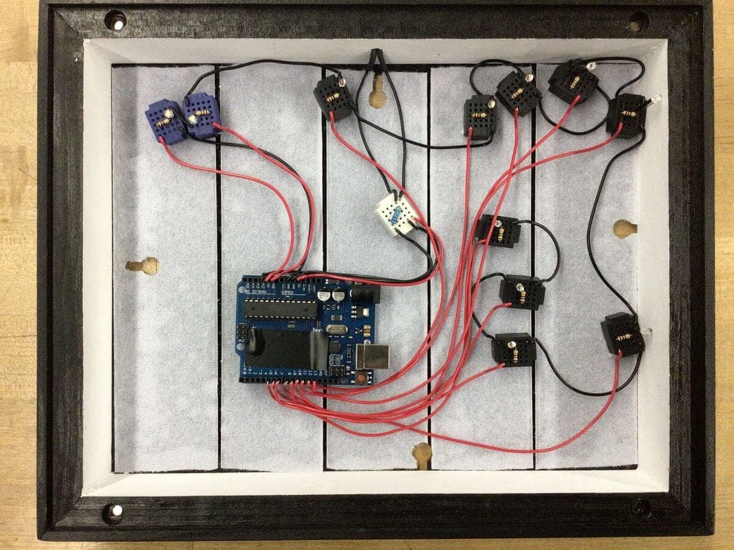 Top-down view into a black frame with white inner walls: a blue Arduino wired with red and black leads to small black breadboards holding LEDs.