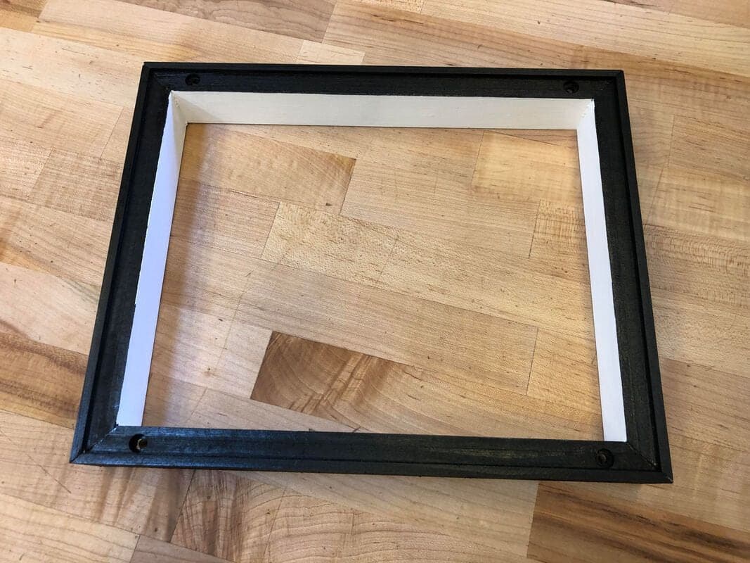 Empty rectangular wooden frame, black outside and white inside, lying on a butcher-block bench with a small drilled hole near each corner.