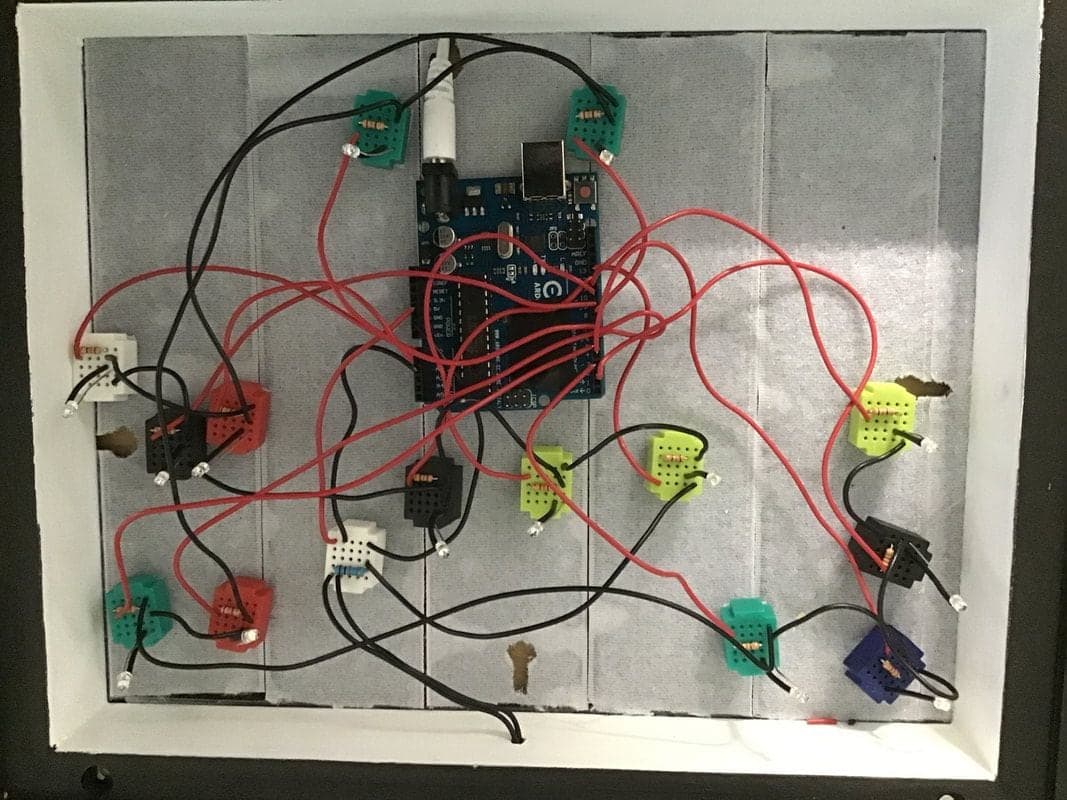Inside a white-walled frame, an Arduino board with red and black wires running to many small coloured breadboards, each holding an LED and resistor.