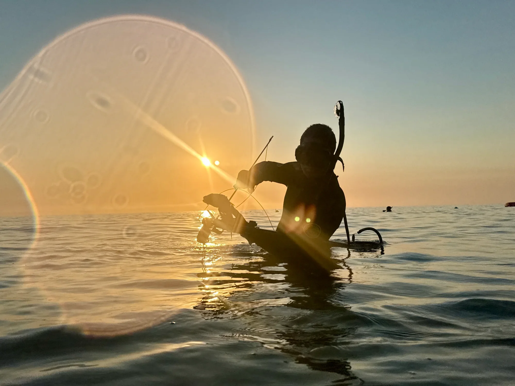 A spearfisher silhouetted against a golden sunset over the water.