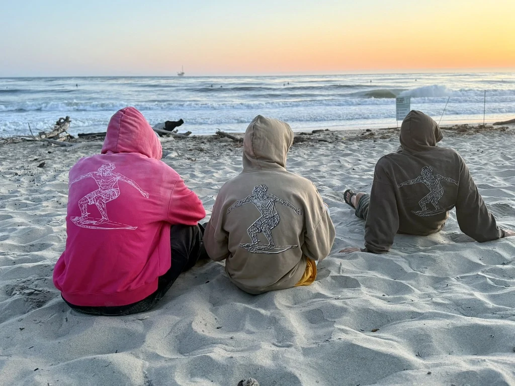 Three friends in 21 Waves hoodies sitting on the sand, watching the surf at sunset.
