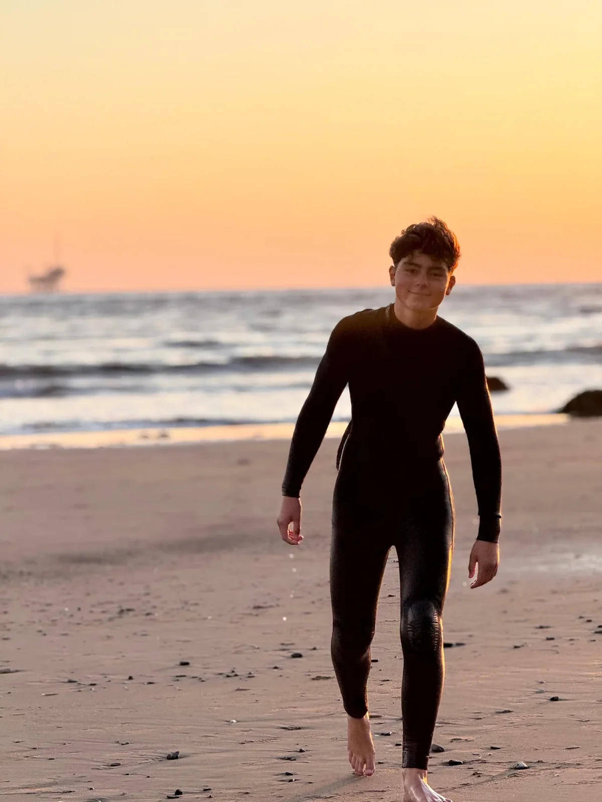 A surfer in a wetsuit walking along the beach at sunset.